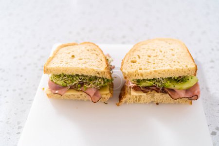 Téléchargez les photos : Assembling ham, cucumber, and sprout sandwiches on the white cutting board. - en image libre de droit