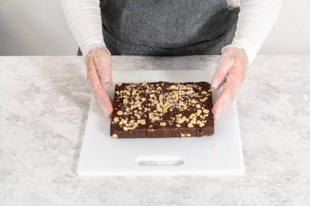 Foto de Corte de caramelo de avellana de chocolate con un cuchillo de cocina grande en trozos cuadrados en una tabla de cortar blanca. - Imagen libre de derechos