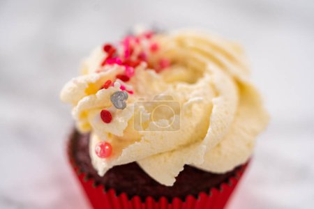 Téléchargez les photos : Piping glaçage ganache chocolat blanc sur le dessus de cupcakes en velours rouge et garniture de saupoudrer. - en image libre de droit