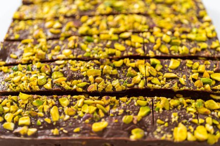 Photo for Cutting chocolate pistachio fudge with a large kitchen knife into square pieces on a white cutting board. - Royalty Free Image