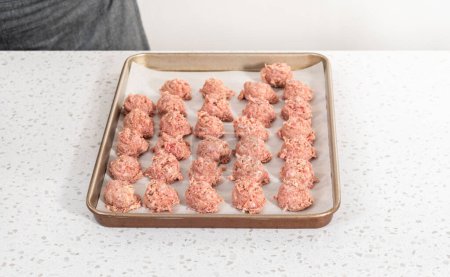 Téléchargez les photos : Ramasser la viande hachée avec une pelle à pâte dans une plaque à pâtisserie tapissée de papier parchemin pour préparer des boulettes de viande cuites au four. - en image libre de droit
