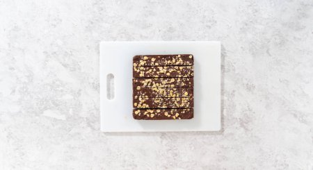 Foto de Acostado. Corte de caramelo de avellana de chocolate con un cuchillo de cocina grande en trozos cuadrados en una tabla de cortar blanca. - Imagen libre de derechos