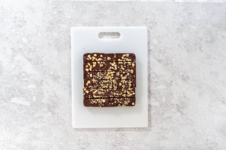 Photo for Flat lay. Cutting chocolate hazelnut fudge with a large kitchen knife into square pieces on a white cutting board. - Royalty Free Image