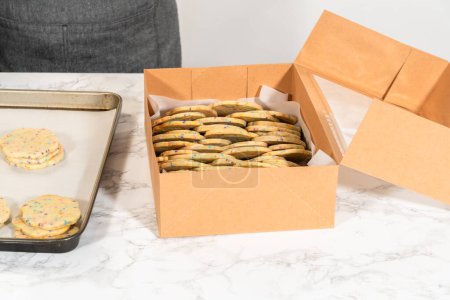 Foto de Con precisión, la mujer está organizando cuidadosamente las galletas de azúcar, llenas de aspersiones mezcladas con masa, en una caja de papel marrón rústico. - Imagen libre de derechos