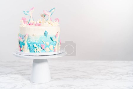 Photo for Mermaid-themed 3 layer vanilla cake decorated with chocolate mermaid tails and seashells on a white cake stand. - Royalty Free Image