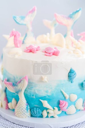 Téléchargez les photos : Gâteau à la vanille 3 couches sur le thème de la sirène décoré de queues de sirène au chocolat et de coquillages sur un stand de gâteau blanc. - en image libre de droit
