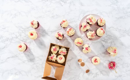 Téléchargez les photos : Pose plate. Emballage fraîchement cuit cupcakes en velours rouge avec glaçage ganache au chocolat blanc dans la boîte à cupcakes. - en image libre de droit