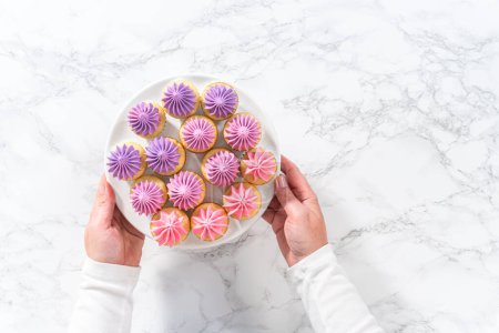 Foto de Acostado. Mini cupcakes de vainilla recién horneados con glaseado de crema de mantequilla rosa ombre en un soporte de pastel blanco. - Imagen libre de derechos