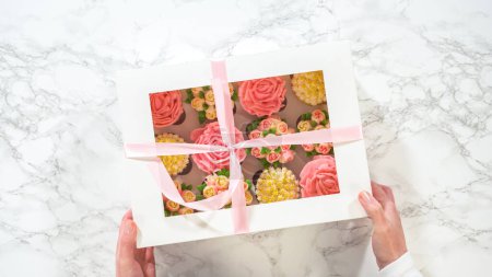 Téléchargez les photos : Pose plate. Tenue par une femme, une boîte en papier blanc révèle des cupcakes gourmands, joliment décorés de glaçage à la crème au beurre façonné en roses et tulipes vibrantes, un témoignage de la créativité culinaire. - en image libre de droit