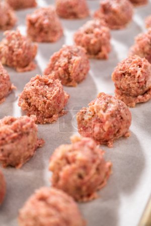 Téléchargez les photos : Ramasser la viande hachée avec une pelle à pâte dans une plaque à pâtisserie tapissée de papier parchemin pour préparer des boulettes de viande cuites au four. - en image libre de droit