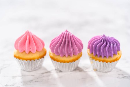 Foto de Mini cupcakes de vainilla recién horneados con glaseado de crema de mantequilla rosa ombre en un ocunter de cocina de mármol. - Imagen libre de derechos