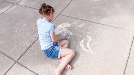 Téléchargez les photos : Petite fille dessinant de l'art crayeux sur une allée de banlieue un jour d'été. - en image libre de droit