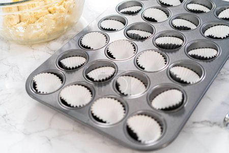 Téléchargez les photos : Scooping pâte cupcake avec cuillère à pâte dans une casserole cupcake cuisson avec doublures pour cuire mini cupcakes vanille avec ombre rose crème au beurre glaçage. - en image libre de droit
