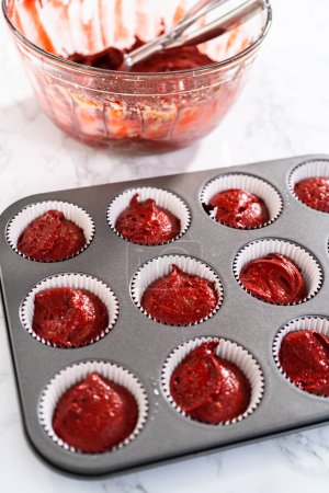 Téléchargez les photos : Scooping pâte cupcake avec pelle à pâte dans une casserole à cupcake doublée de doublures en papier d'aluminium pour cuire des cupcakes en velours rouge avec glaçage ganache au chocolat blanc. - en image libre de droit