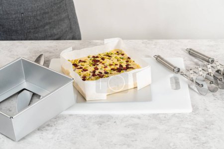Photo for Removing cranberry pistachio fudge from a square cheesecake pan lined with parchment. - Royalty Free Image
