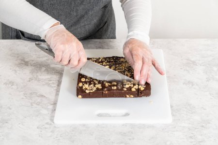 Téléchargez les photos : Découpe de caramel aux noisettes au chocolat avec un grand couteau de cuisine en morceaux carrés sur une planche à découper blanche. - en image libre de droit
