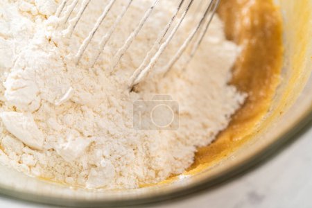 Photo for Mixing wet and dry ingredients with a hand whisk in a glass mixing bowl to bake white chocolate macadamia nut. - Royalty Free Image