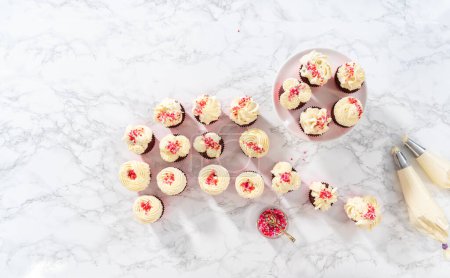 Photo for Flat lay. Piping white chocolate ganache frosting on top of red velvet cupcakes and topping with sprinkles. - Royalty Free Image