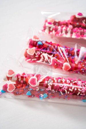 Barras de pretzel cubiertas de chocolate decoradas con aspersiones en forma de corazón para el Día de San Valentín empaquetadas en bolsas transparentes.