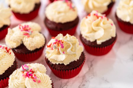 Foto de Piping glaseado de ganache de chocolate blanco en la parte superior de cupcakes de terciopelo rojo y cubierta con aspersiones. - Imagen libre de derechos