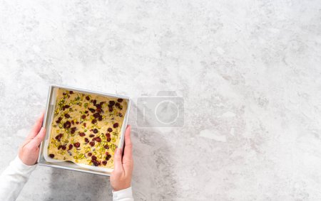 Téléchargez les photos : Pose plate. Poêle à gâteau au fromage carrée garnie de papier sulfurisé avec mélange de caramel pour préparer le caramel à la pistache aux canneberges. - en image libre de droit