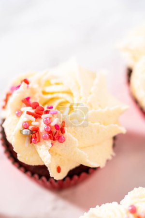 Photo for Piping white chocolate ganache frosting on top of red velvet cupcakes and topping with sprinkles. - Royalty Free Image