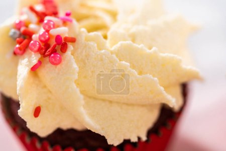 Photo for Piping white chocolate ganache frosting on top of red velvet cupcakes and topping with sprinkles. - Royalty Free Image