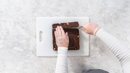 Photo for Flat lay. Cutting chocolate macadamia fudge with a large kitchen knife into square pieces on a white cutting board. - Royalty Free Image
