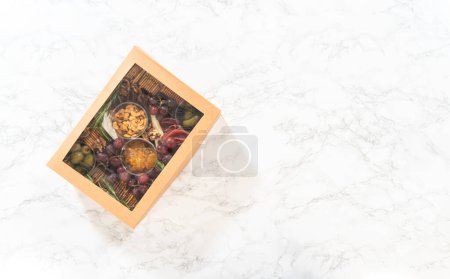 Téléchargez les photos : Pose plate. Boîte de charcuterie avec viande tranchée, fromage, craquelins et raisins, le tout soigneusement emballé dans une boîte à cadeaux marron. - en image libre de droit
