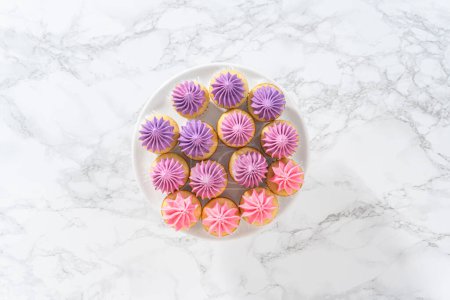 Foto de Acostado. Mini cupcakes de vainilla recién horneados con glaseado de crema de mantequilla rosa ombre en un soporte de pastel blanco. - Imagen libre de derechos