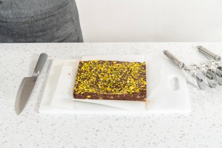 Téléchargez les photos : Removing chocolate pistachio fudge from a square cheesecake pan lined with parchment. - en image libre de droit
