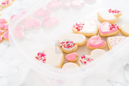 Foto de Almacenamiento de galletas de azúcar en forma de corazón con glaseado real rosa y blanco en un recipiente de plástico grande. - Imagen libre de derechos