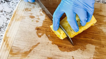 Téléchargez les photos : Portant des gants jetables bleus pour l'hygiène, quelqu'un tranche habilement dans une pastèque bio juteuse sur une planche à découper en bois, posée sur un comptoir de cuisine propre. - en image libre de droit