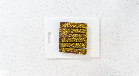 Téléchargez les photos : Pose plate. Découpe de caramel au chocolat à la pistache avec un grand couteau de cuisine en morceaux carrés sur une planche à découper blanche. - en image libre de droit