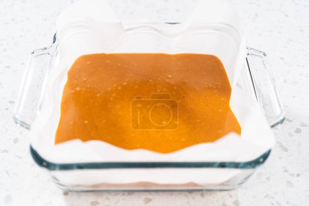 Téléchargez les photos : Verser le mélange de caramel dans le moule en verre carré doublé de papier parchemin pour préparer le caramel à la citrouille. - en image libre de droit