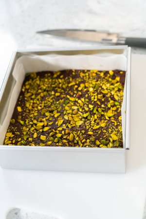 Téléchargez les photos : Removing chocolate pistachio fudge from a square cheesecake pan lined with parchment. - en image libre de droit