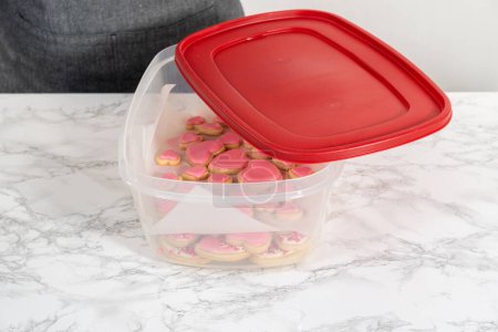 Aufbewahrung herzförmiger Zuckerkekse mit rosa und weißer Zuckerglasur in einem großen Plastikbehälter.