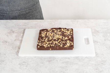 Foto de Corte de caramelo de avellana de chocolate con un cuchillo de cocina grande en trozos cuadrados en una tabla de cortar blanca. - Imagen libre de derechos