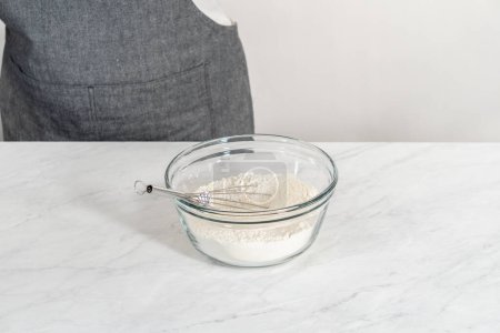Foto de Mezclar ingredientes secos con un batidor de mano en un tazón de vidrio para hornear nuez de macadamia de chocolate blanco. - Imagen libre de derechos