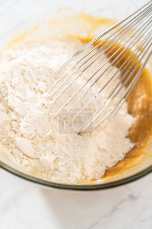 Photo for Mixing wet and dry ingredients with a hand whisk in a glass mixing bowl to bake white chocolate macadamia nut. - Royalty Free Image