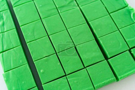 Téléchargez les photos : Couper le caramel vert avec un grand couteau de cuisine en morceaux carrés sur une planche à découper blanche. - en image libre de droit