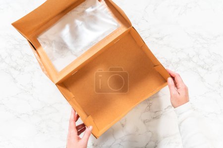 Photo for Flat lay. In the process of assembling, a woman expertly curates this charcuterie box, showcasing sliced meat, cheese, crackers, and grapes, all neatly arranged and packaged in a brown gifting box. - Royalty Free Image