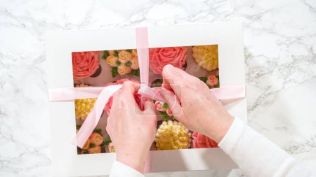 Foto de Acostado. En las manos de una mujer, una caja de papel blanco llena de cupcakes gourmet, cada uno adornado con rosas vibrantes y tulipanes hechos a mano de glaseado de crema de mantequilla, se está cerrando suavemente. - Imagen libre de derechos