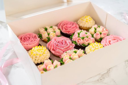 Eingehüllt in eine makellose weiße Papier-Cupcake-Box ist jeder Gourmet-Cupcake ein Kunstwerk, geschmückt mit Buttercreme-Zuckerguss-Blüten, die wunderschön gestaltet sind, um lebendigen Rosen und Tulpen zu ähneln.