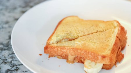 In the stylish ambiance of a modern white kitchen, a grilled peanut butter and banana sandwich is expertly sliced and tastefully presented on a pristine white plate.