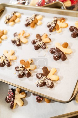Téléchargez les photos : Création de biscuits sucrés découpés, partiellement trempés dans du chocolat et garnis de morceaux de noisettes, placés sur du papier parchemin. - en image libre de droit