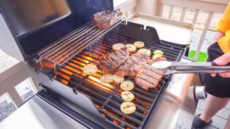 Téléchargez les photos : Le barbecue à gaz extérieur à deux brûleurs est mis à bon usage, pétillant avec le son et l'arôme des steaks de côtelette et des rondelles d'oignon étant parfaitement cuits. - en image libre de droit