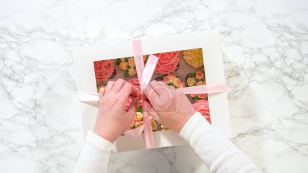 Foto de Acostado. Las manos de una mujer abren suavemente una caja de papel blanco, revelando un conjunto de cupcakes gourmet en el interior, cada uno artísticamente adornado con glaseado de crema de mantequilla, diseñado para imitar vibrantes rosas y tulipanes. - Imagen libre de derechos