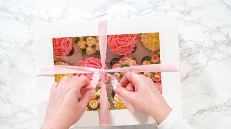 Téléchargez les photos : Pose plate. Dans une main de femme, une boîte en papier blanc remplie de cupcakes gourmands, chacun orné de roses vibrantes et de tulipes fabriquées à partir de glaçage à la crème au beurre, est doucement fermée. - en image libre de droit