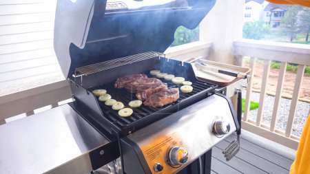 Téléchargez les photos : Le barbecue à gaz extérieur à deux brûleurs est mis à bon usage, pétillant avec le son et l'arôme des steaks de côtelette et des rondelles d'oignon étant parfaitement cuits. - en image libre de droit